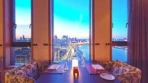 Un comedor con una mesa dispuesta para dos personas, ventanas amplias con vistas a la ciudad y una vela decorativa sobre la mesa.