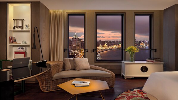 Habitación de hotel con vista al horizonte de la ciudad, un sofá, un escritorio con una computadora portátil y una mesita con libros.
