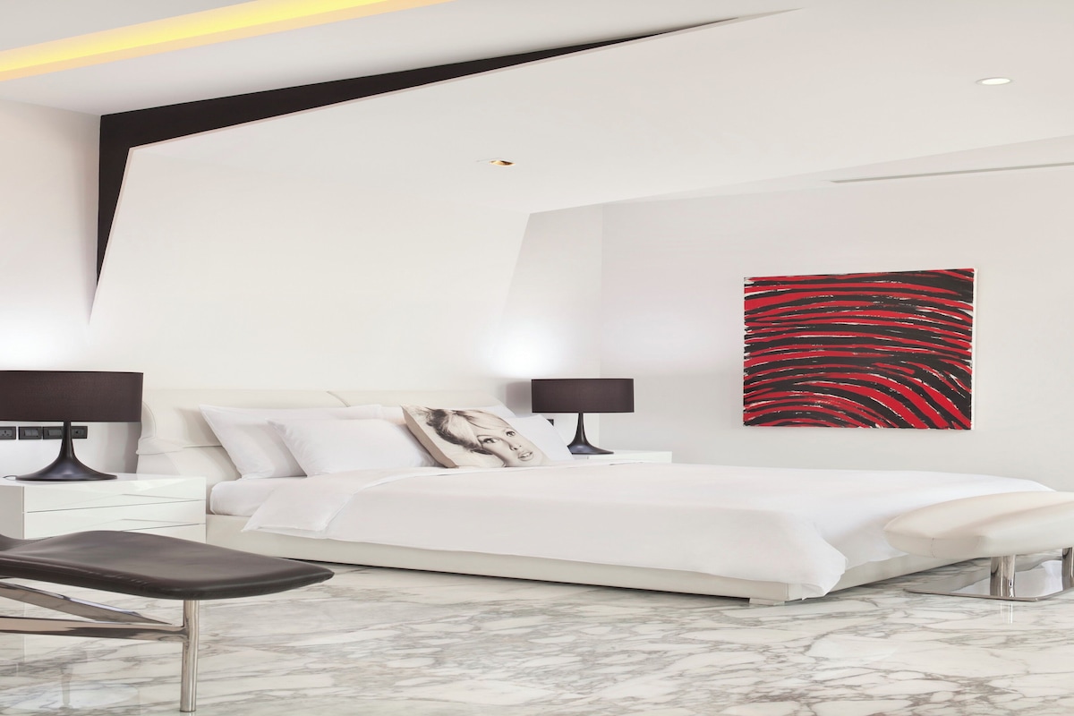 Une chambre à coucher moderne, avec un lit, deux tables de chevet, une chaise et un grand tableau abstrait accroché au mur.