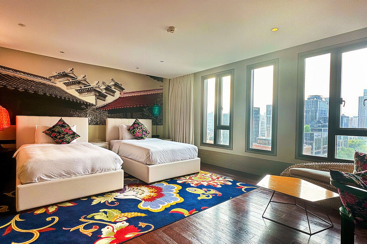 Une chambre d’hôtel avec deux lits, une grande fenêtre donnant sur la ville, un mur décoratif et une moquette à motifs.