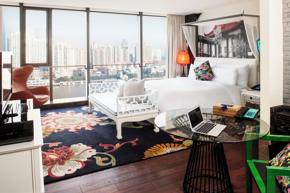 Une chambre d’hôtel moderne avec une grande fenêtre, un lit, un bureau avec un ordinateur portable et une chaise verte.