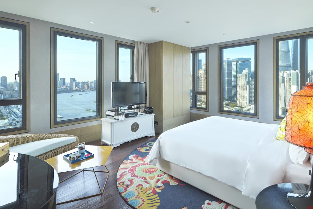 Une chambre d’hôtel avec un grand lit, une télévision, un bureau avec une chaise, un tapis coloré et une vue sur la ville et l’eau, grâce à de grandes fenêtres.