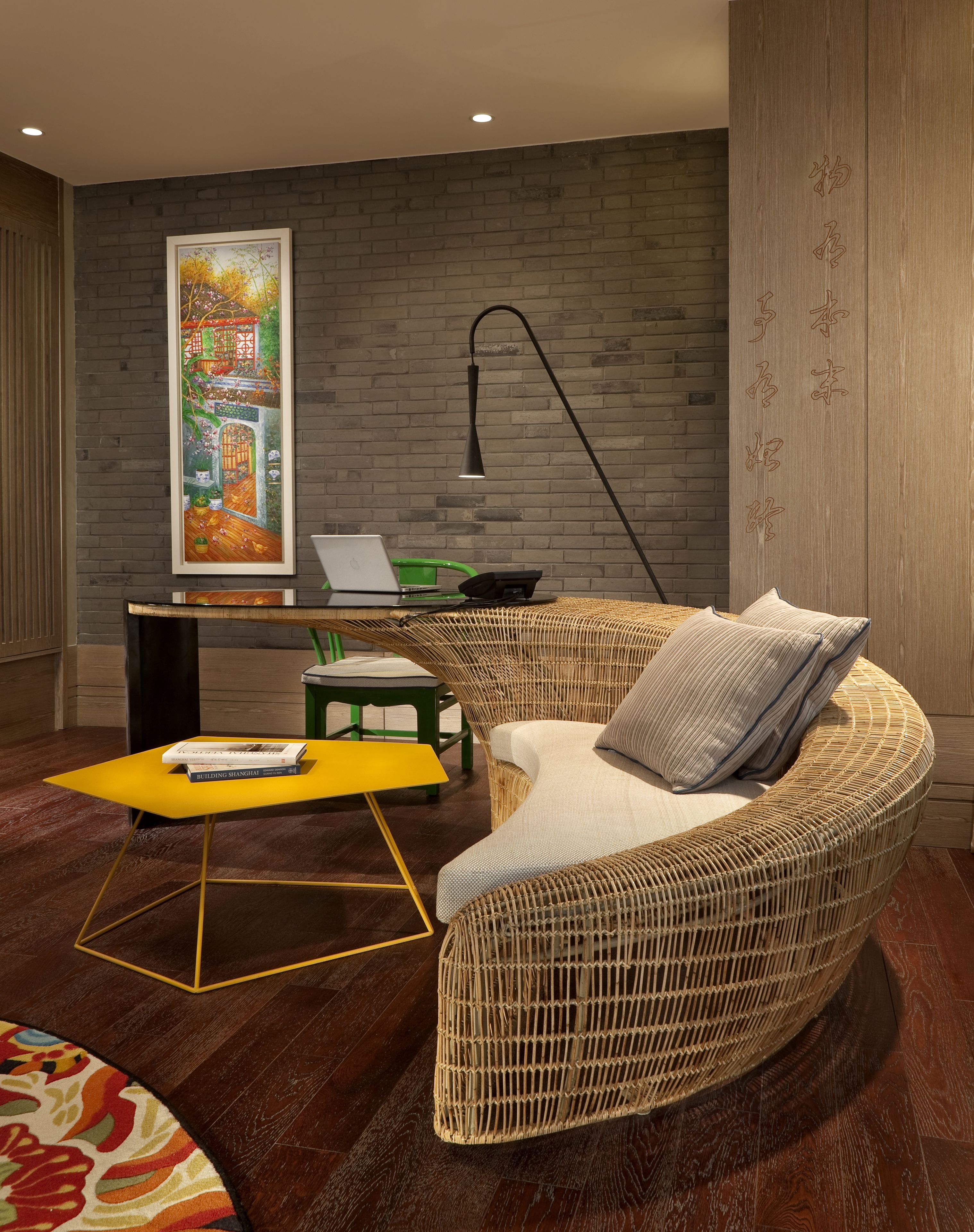 A modern living room with a wicker sofa, a yellow coffee table, a framed painting, and a laptop on a green stool.