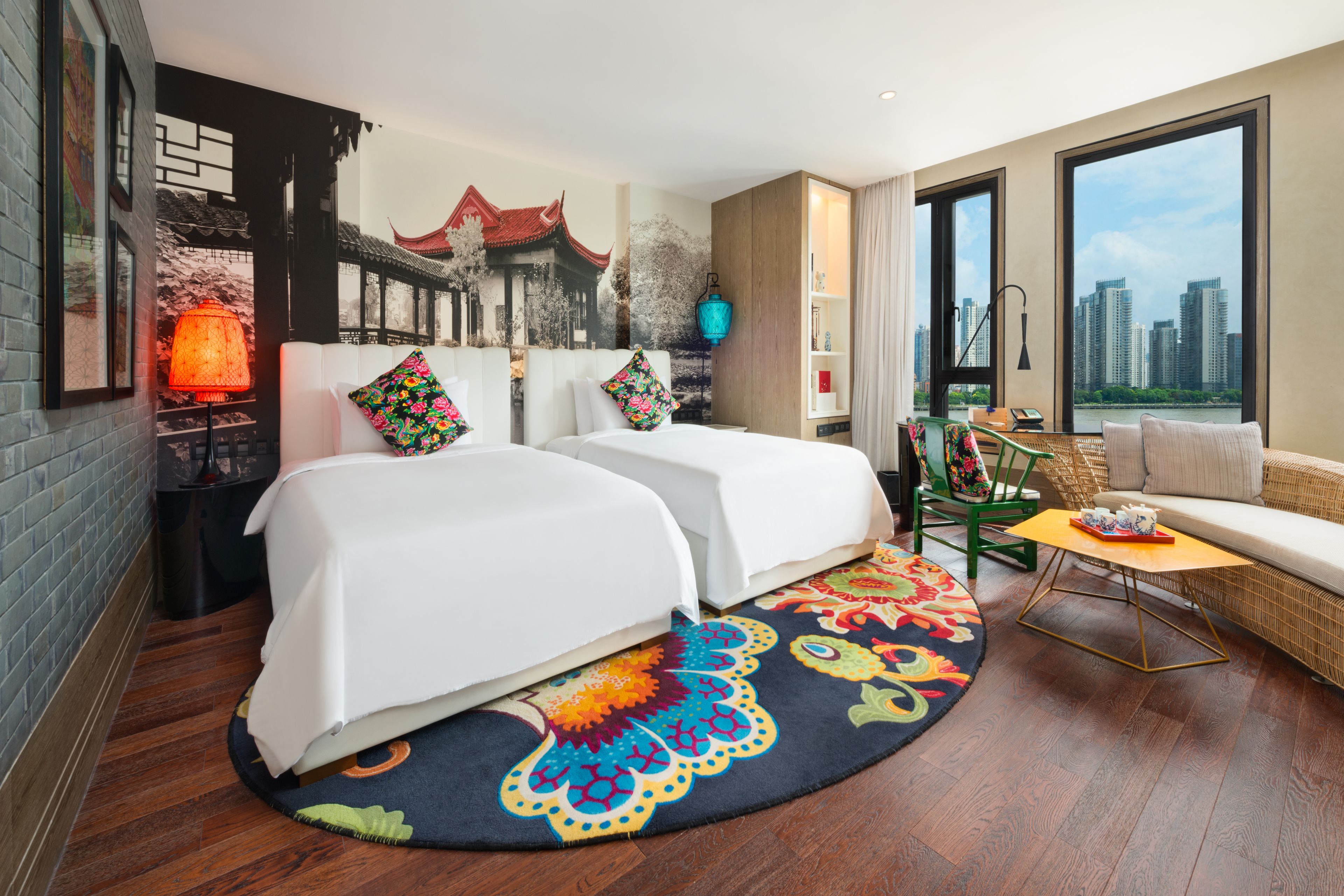 A hotel room with two beds, a sofa, a small table, a colorful rug, and a city view.