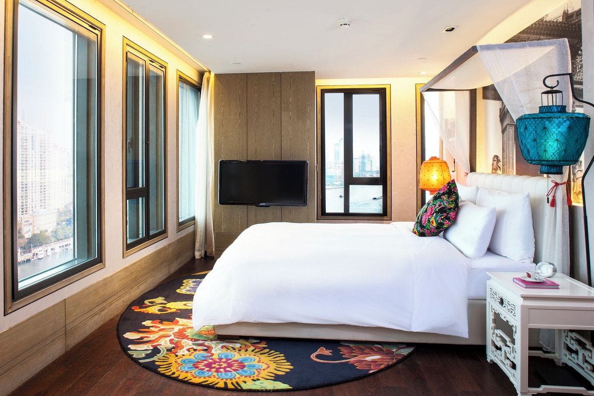 Une chambre d’hôtel avec un grand lit, un téléviseur à écran plat, un tapis aux couleurs vives et une vue sur la ville par les fenêtres.