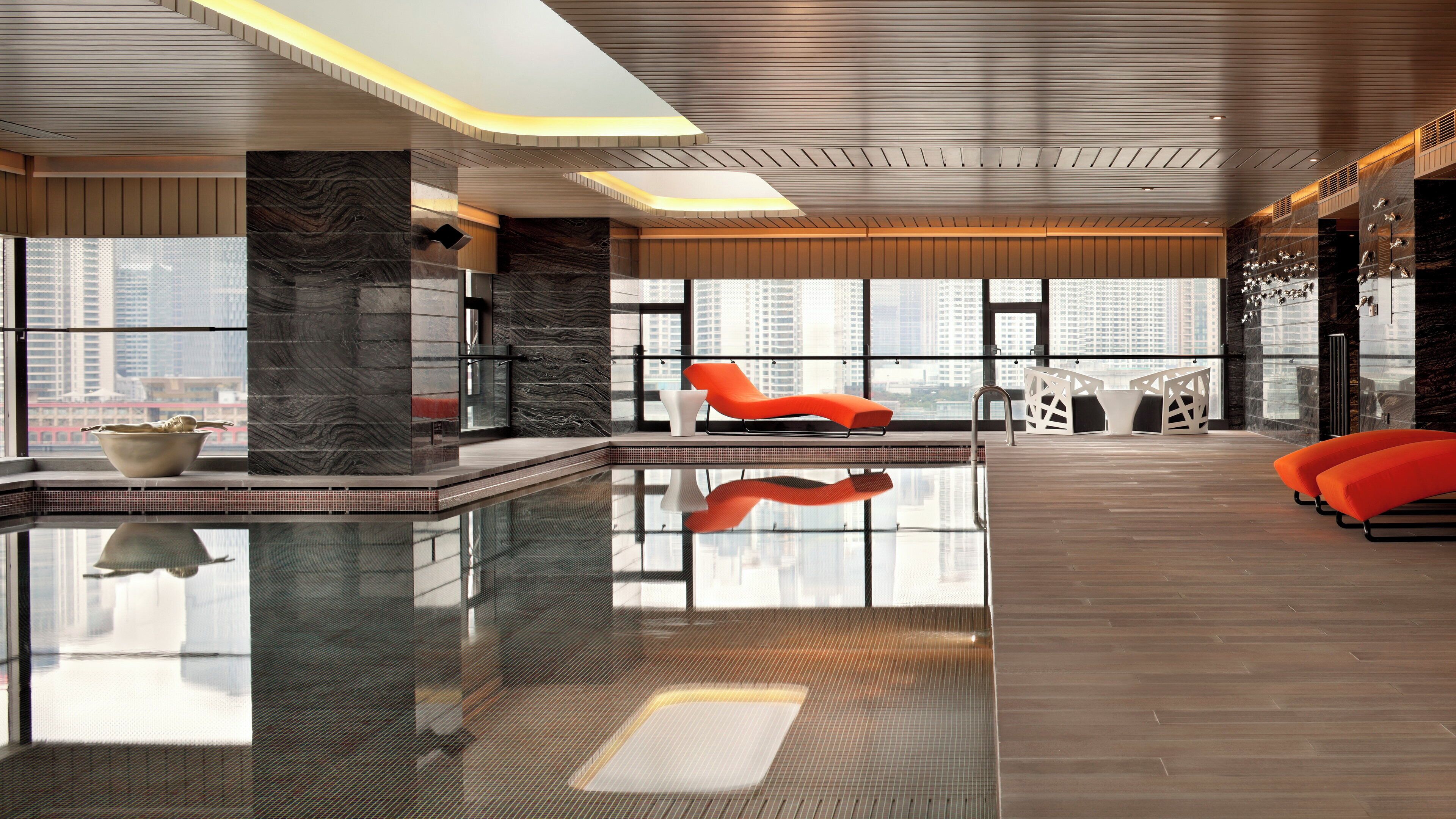 Un área de piscina interior moderna con piso de vidrio, asientos y vistas a la ciudad.