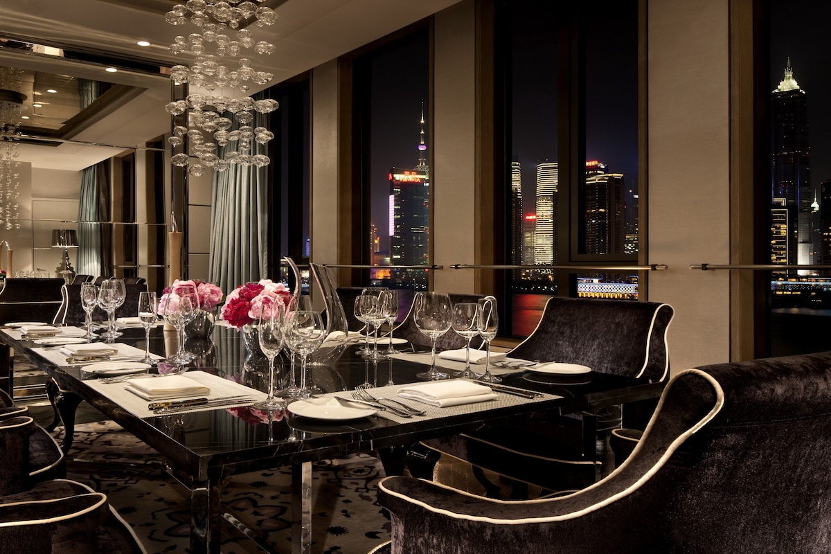 Une salle à manger avec une longue table dressée pour le repas, de grandes fenêtres donnant sur la ville et un lustre.