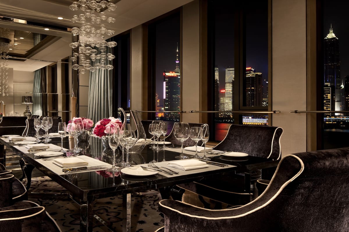 A dining area with a long table set for six, large windows offering a city view, and a modern chandelier.