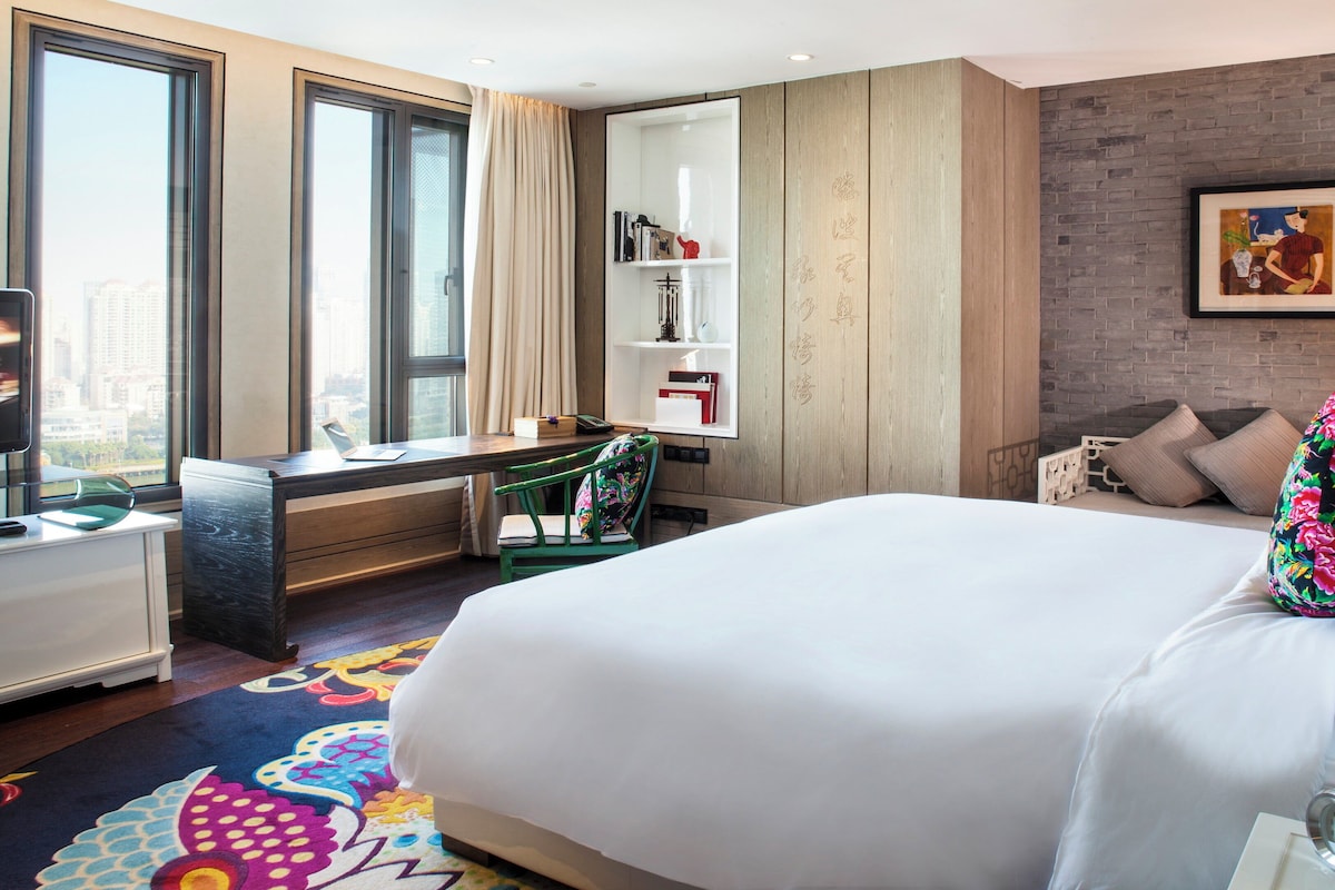 Une chambre d’hôtel avec un lit, un bureau, une chaise, une bibliothèque et une vue sur la ville.