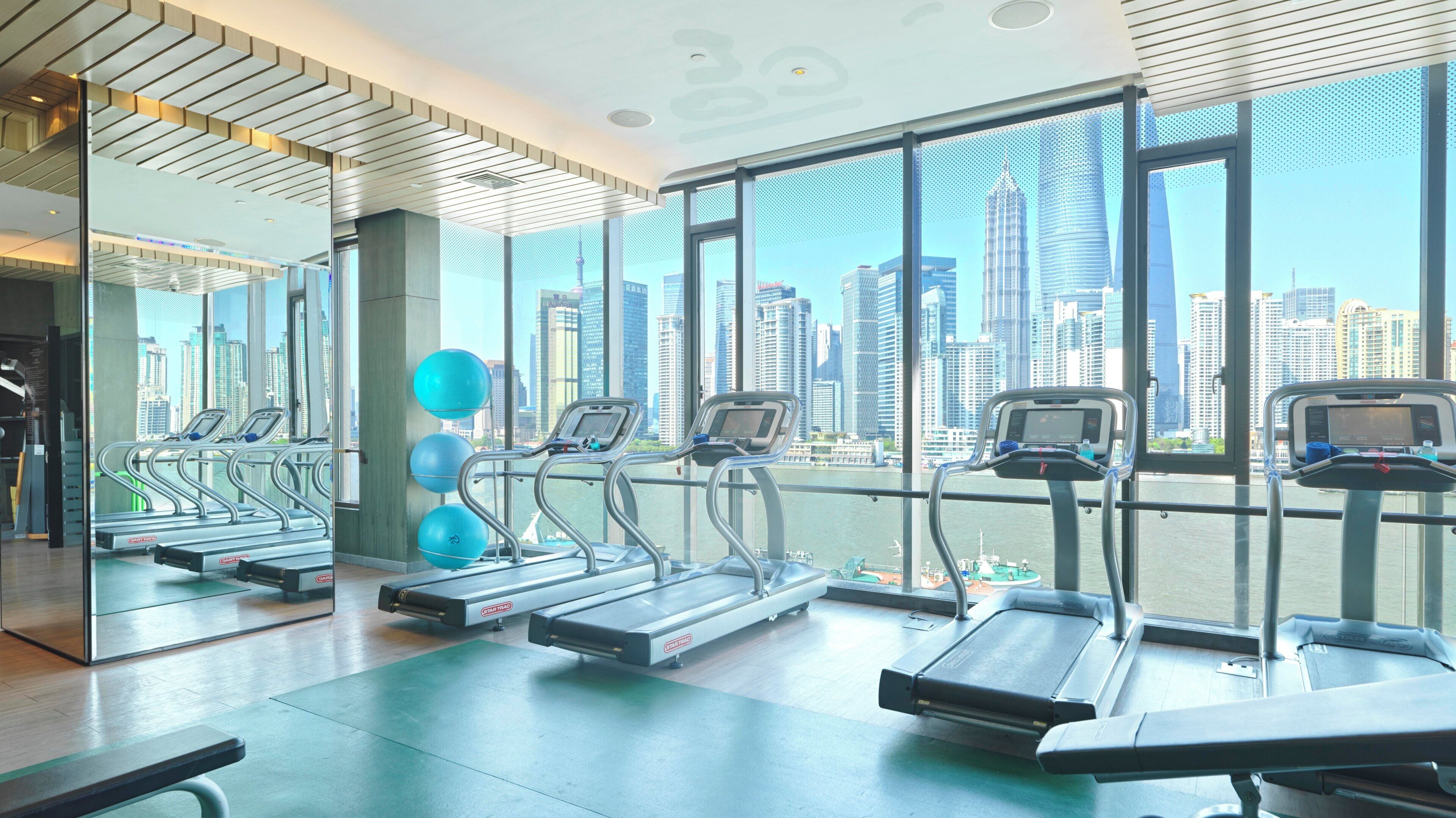A gym with treadmills and exercise balls, large windows with a city view.