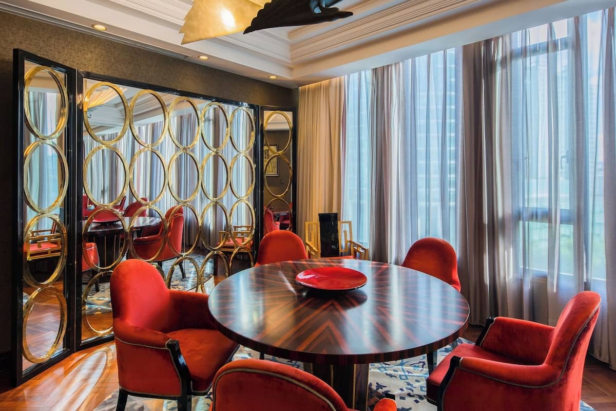 Une chambre d’hôtel avec une table ronde, des chaises rouges, un tapis à motifs, un miroir décoratif et une grande fenêtre avec des rideaux.