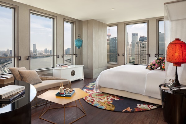Une chambre d’hôtel moderne, dotée d’un grand lit, d’un canapé, d’une petite table et offrant une vue sur le paysage urbain grâce à de grandes fenêtres.