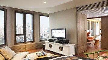 A modern hotel room with large windows, a flat-screen TV, a white console table, a sofa, and a dining area with a tray of items.