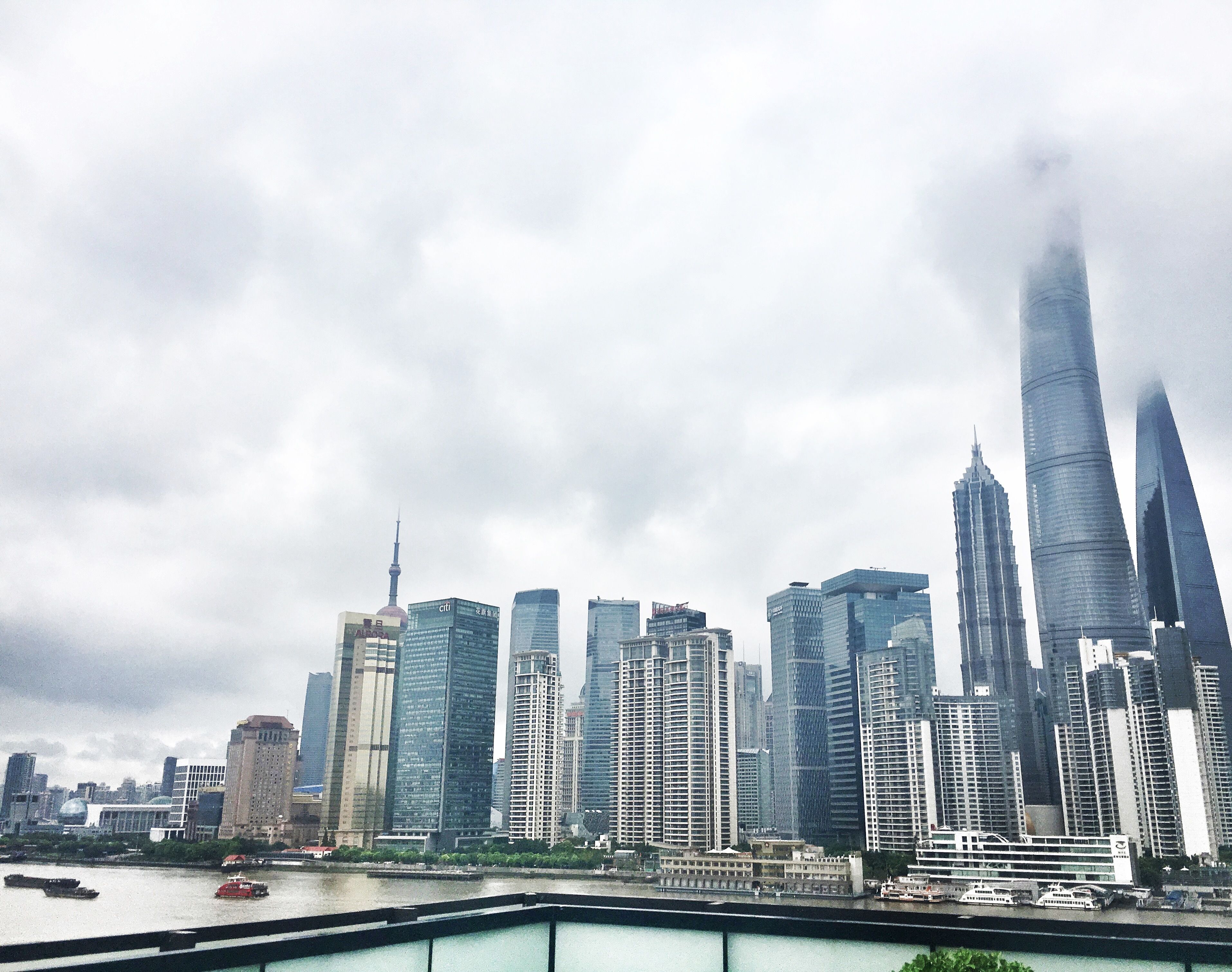 A city skyline with modern high-rise buildings, a river with boats, and a cloudy sky.