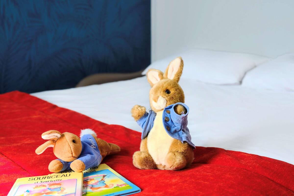 Zwei Plüschtiere liegen auf einem Bett mit einer roten Decke, einem Kissen und einem Buch mit dem Titel 'SOURICEAU et Souricette'.