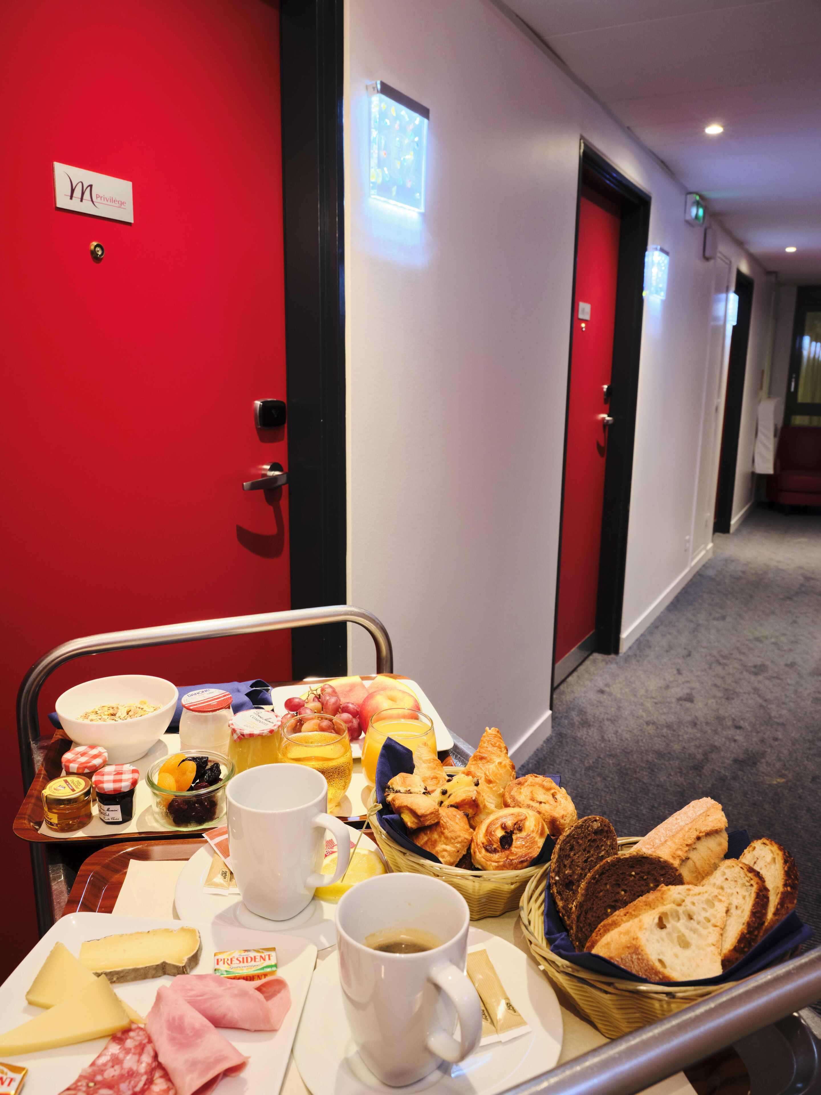 A breakfast trolley with various food items including bread, cheese, and pastries, in a hotel corridor with red doors and a white wall.