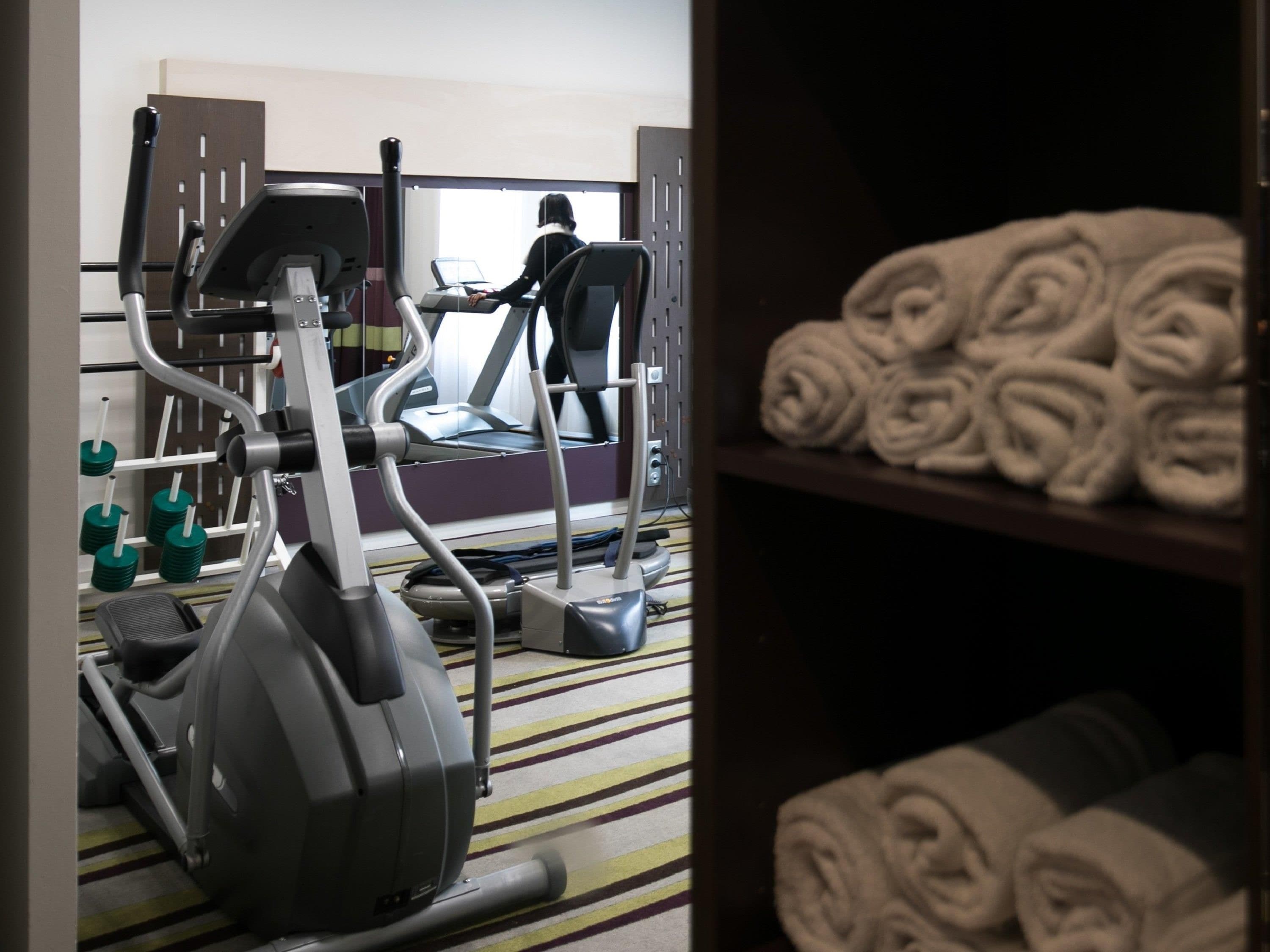 Une salle de sport équipée de matériel d’exercice, d’un tapis de course et de serviettes rangées sur des étagères.