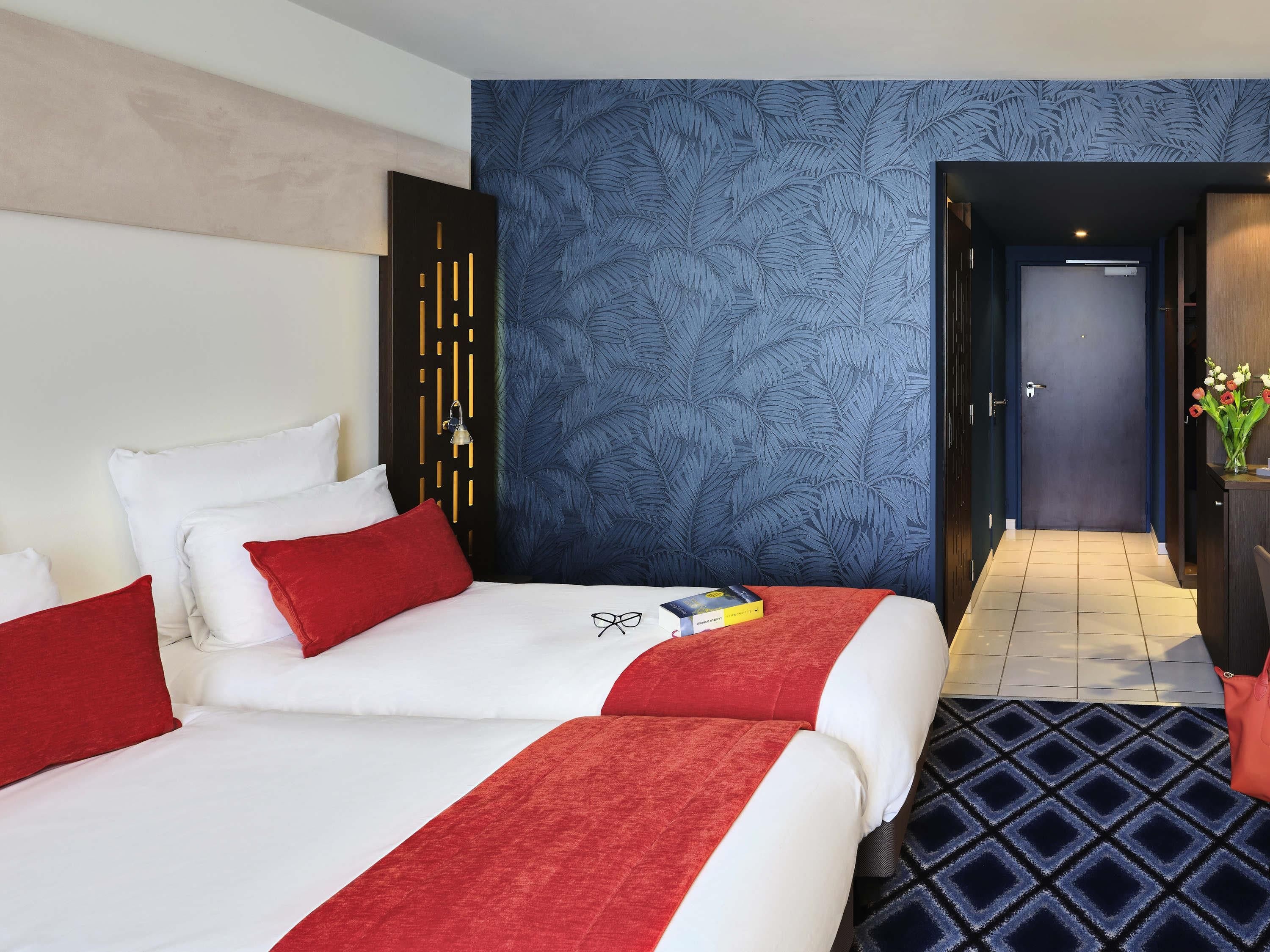A hotel room with two beds, a blue leaf-patterned wall, a dark door, and a tiled floor.