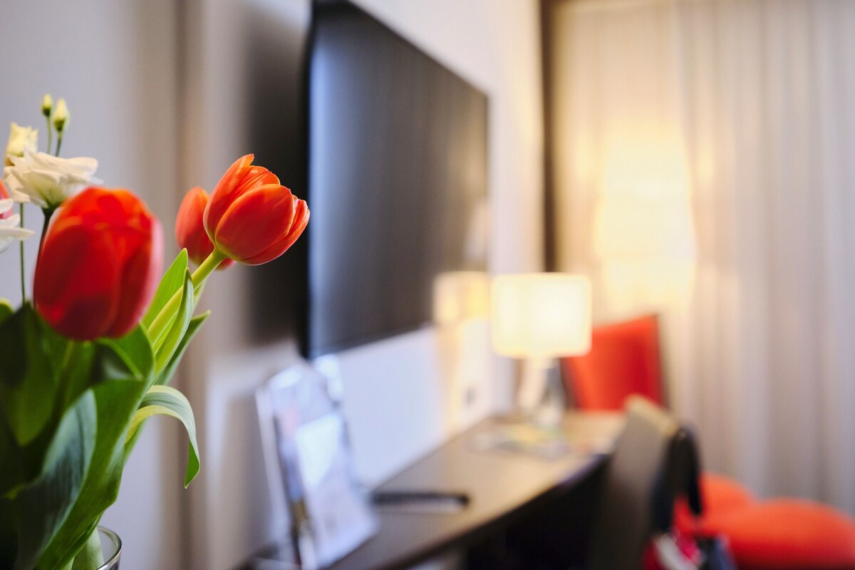 Une chambre d’hôtel avec un lit, un bureau, une chaise, une télévision et un vase avec des fleurs.