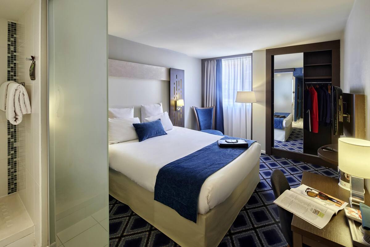 A hotel room with a bed, a desk, a chair, a lamp, a mirror, and a wardrobe.