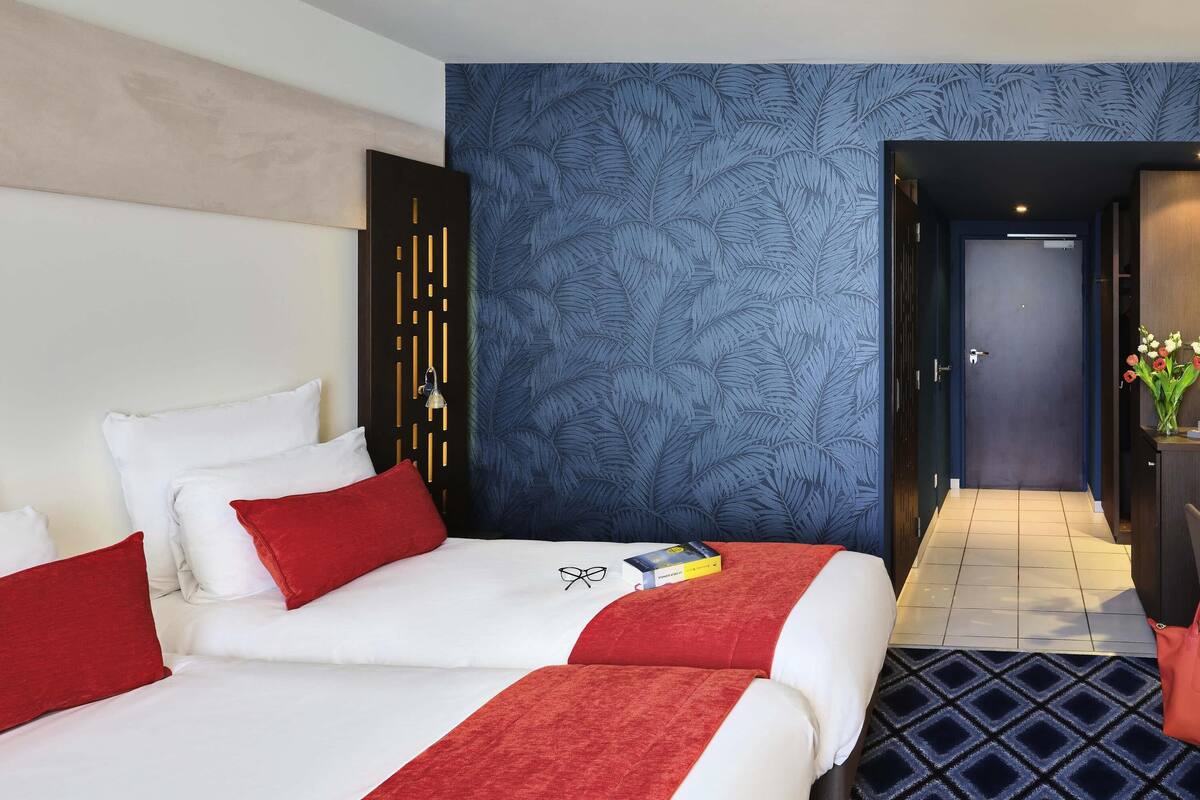 Ein Hotelzimmer mit zwei Betten, einer Wand mit blauem Blattmuster, einer dunklen Tür und einem Fliesenboden.