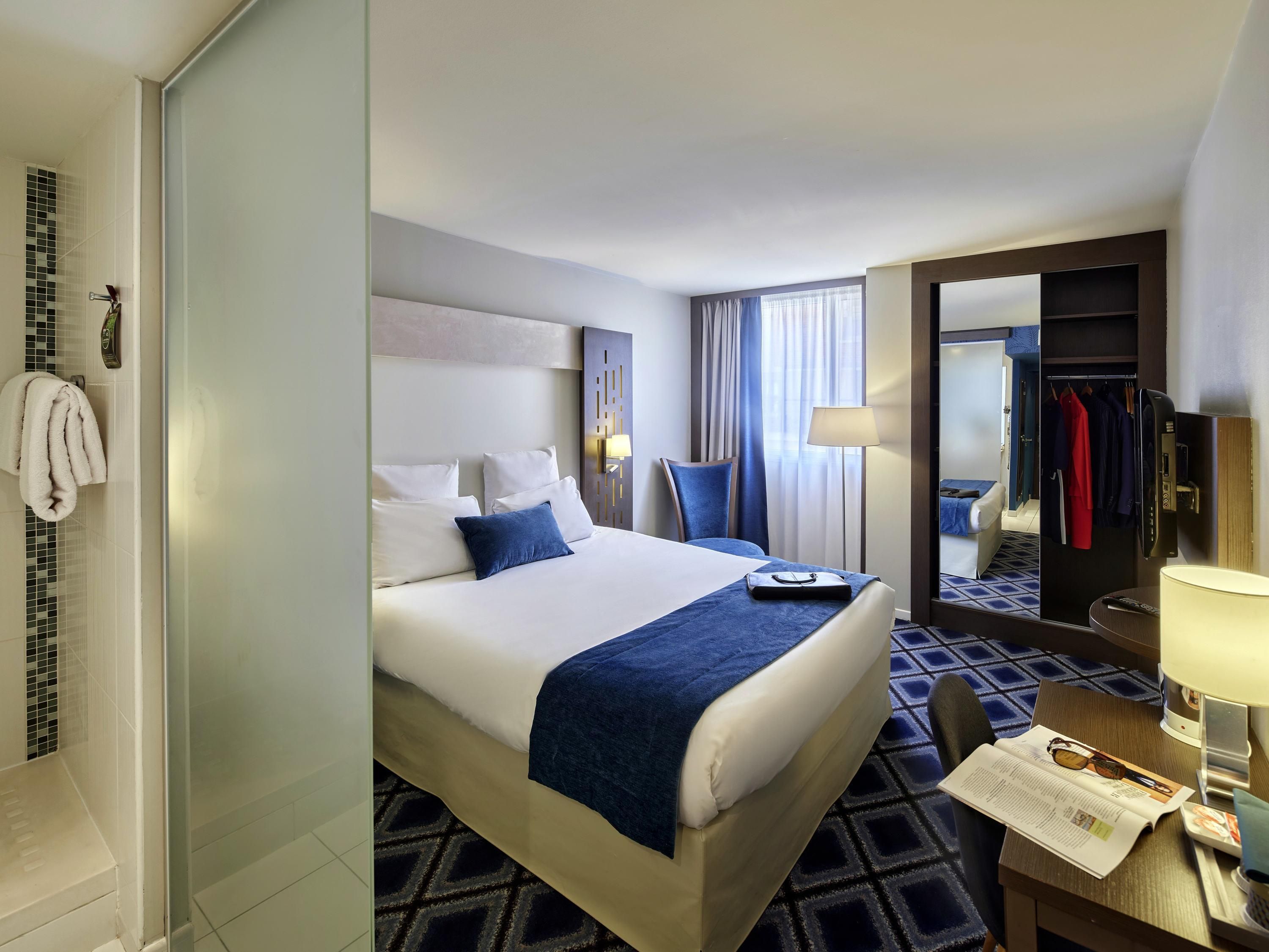 A hotel room with a bed, a desk, a chair, a lamp, a mirror, and a wardrobe.