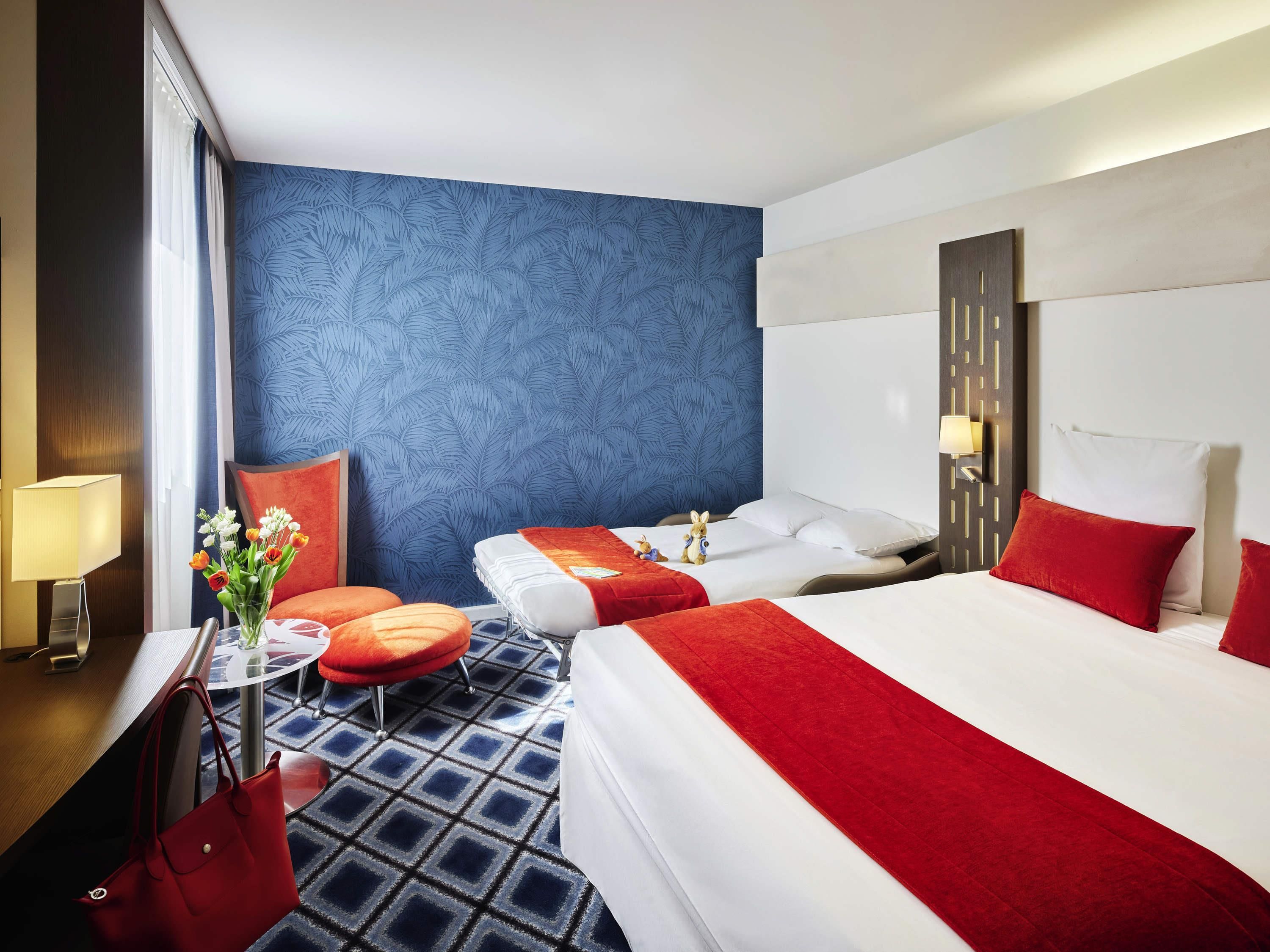 A hotel room with two beds, a red and white color scheme, a blue patterned wall, and a small round table with a lamp.