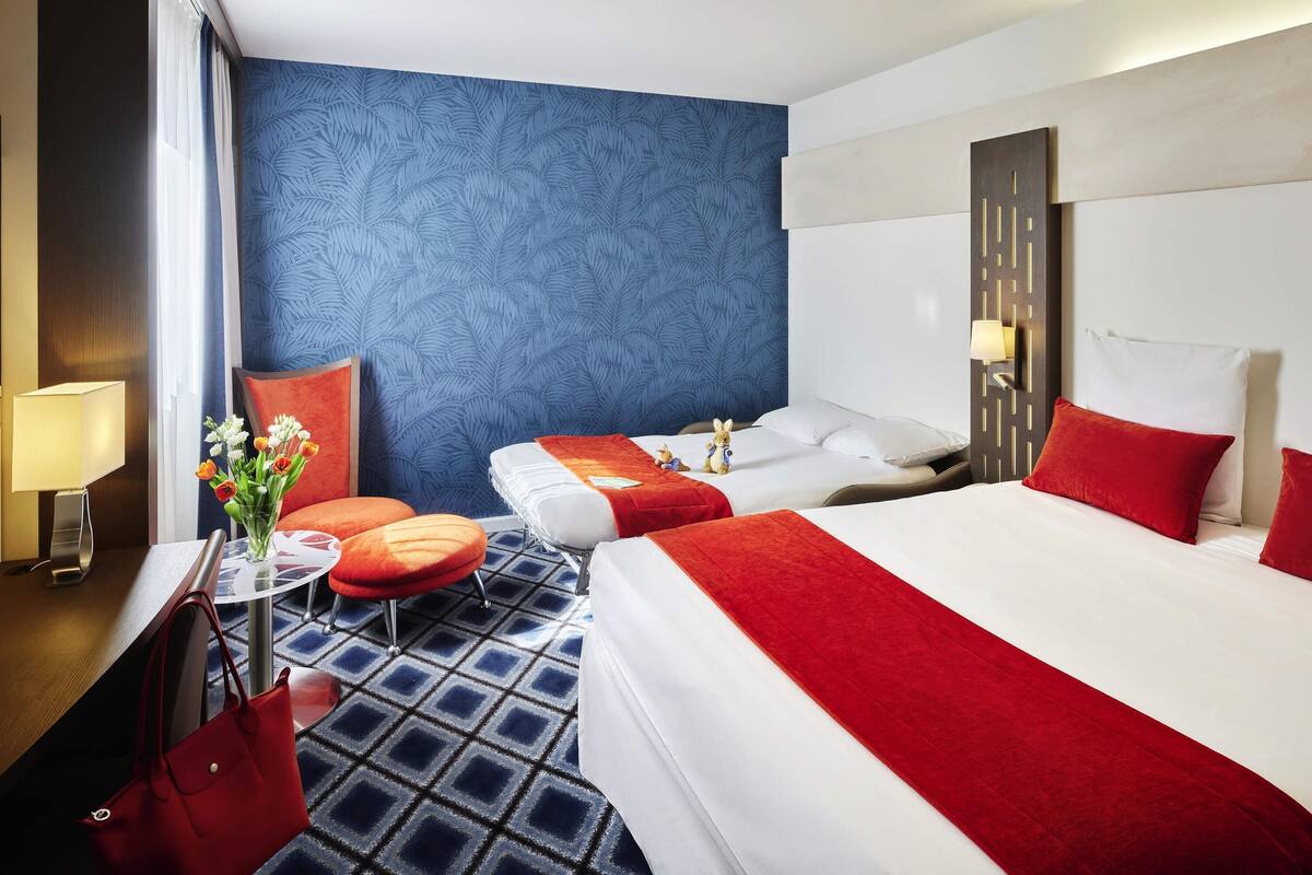 A hotel room with two beds, a red and white color scheme, a blue patterned wall, and a small round table with a lamp.