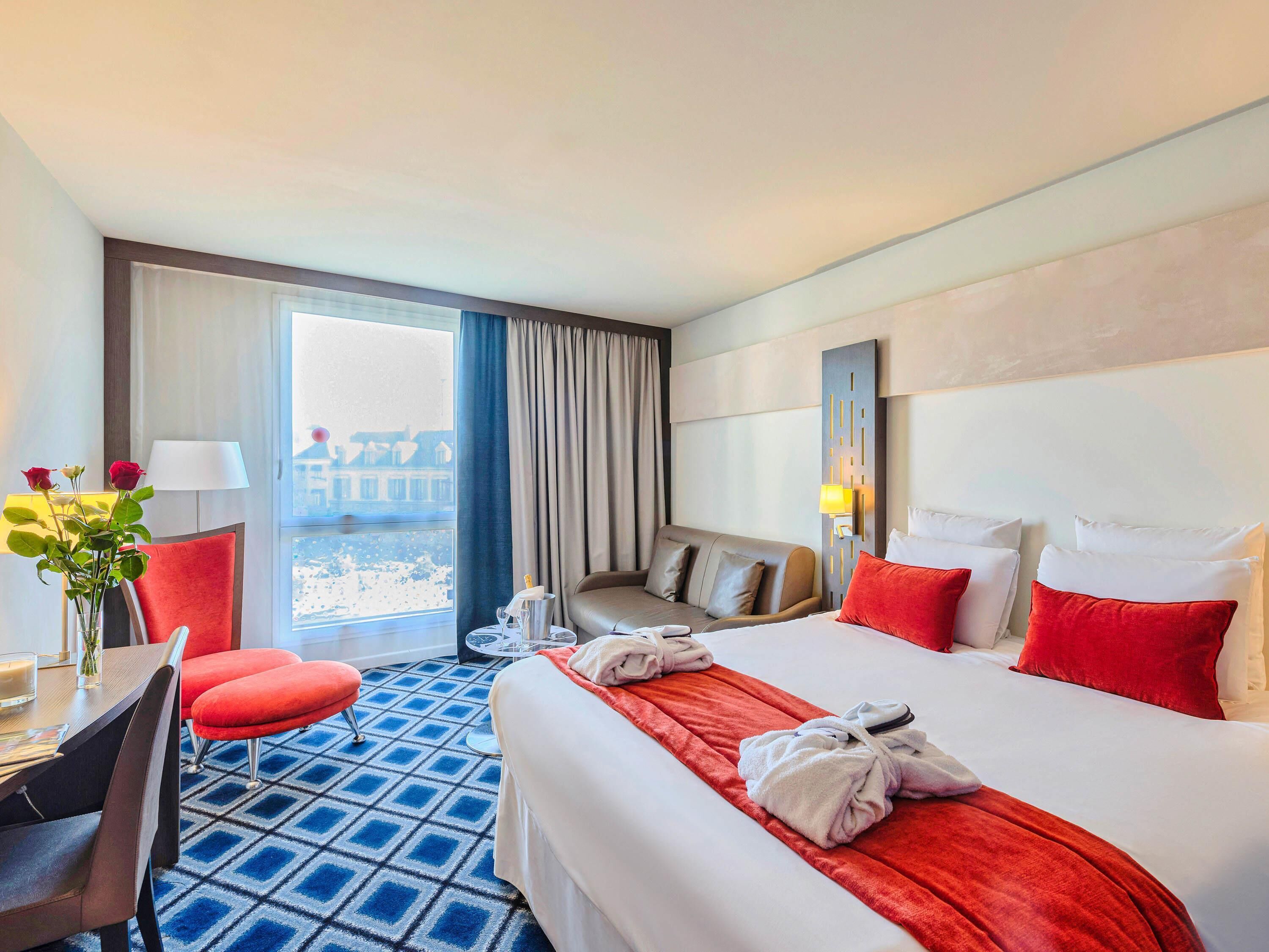 A hotel room with a bed, a desk, a chair, a table, a vase with flowers, and a view of buildings through the window.