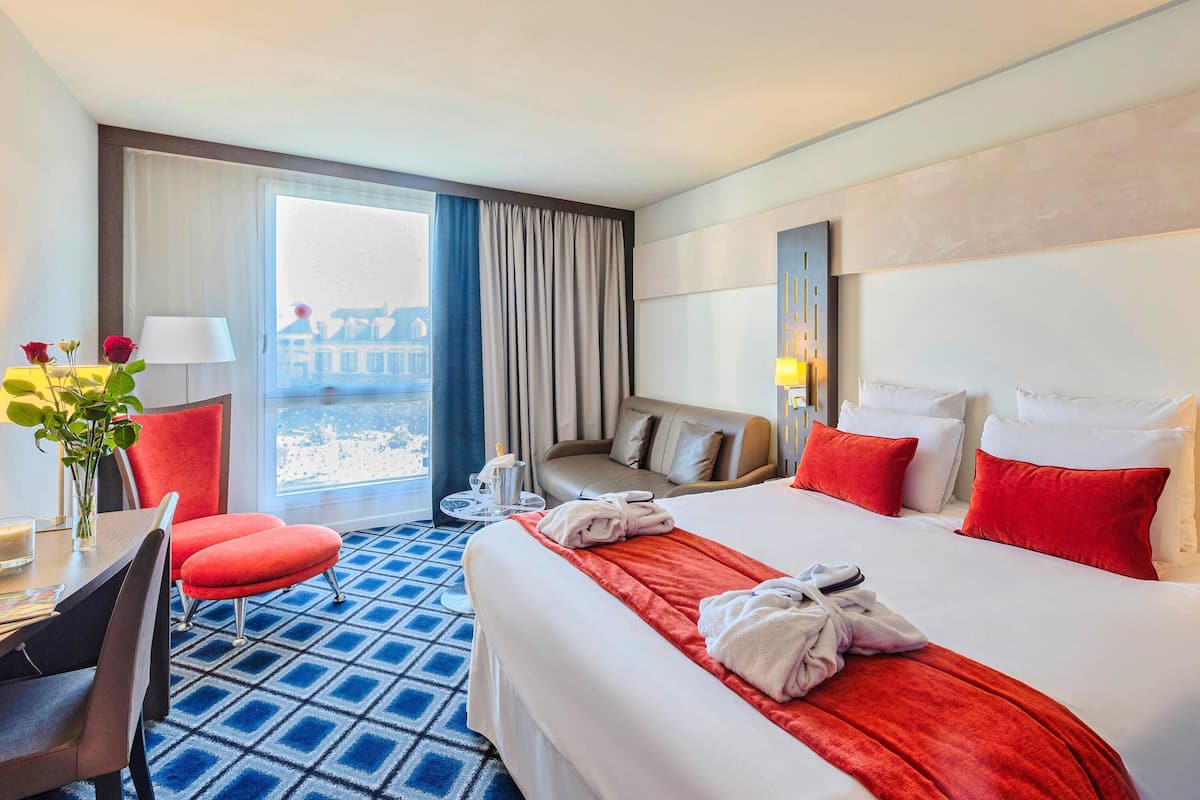 A hotel room with a bed, a desk, a chair, a table, a vase with flowers, and a view of buildings through the window.