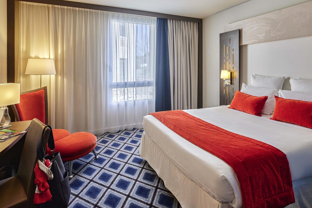 Une chambre d’hôtel avec un grand lit, un bureau avec une chaise, une fenêtre avec des rideaux et un sol à motifs.
