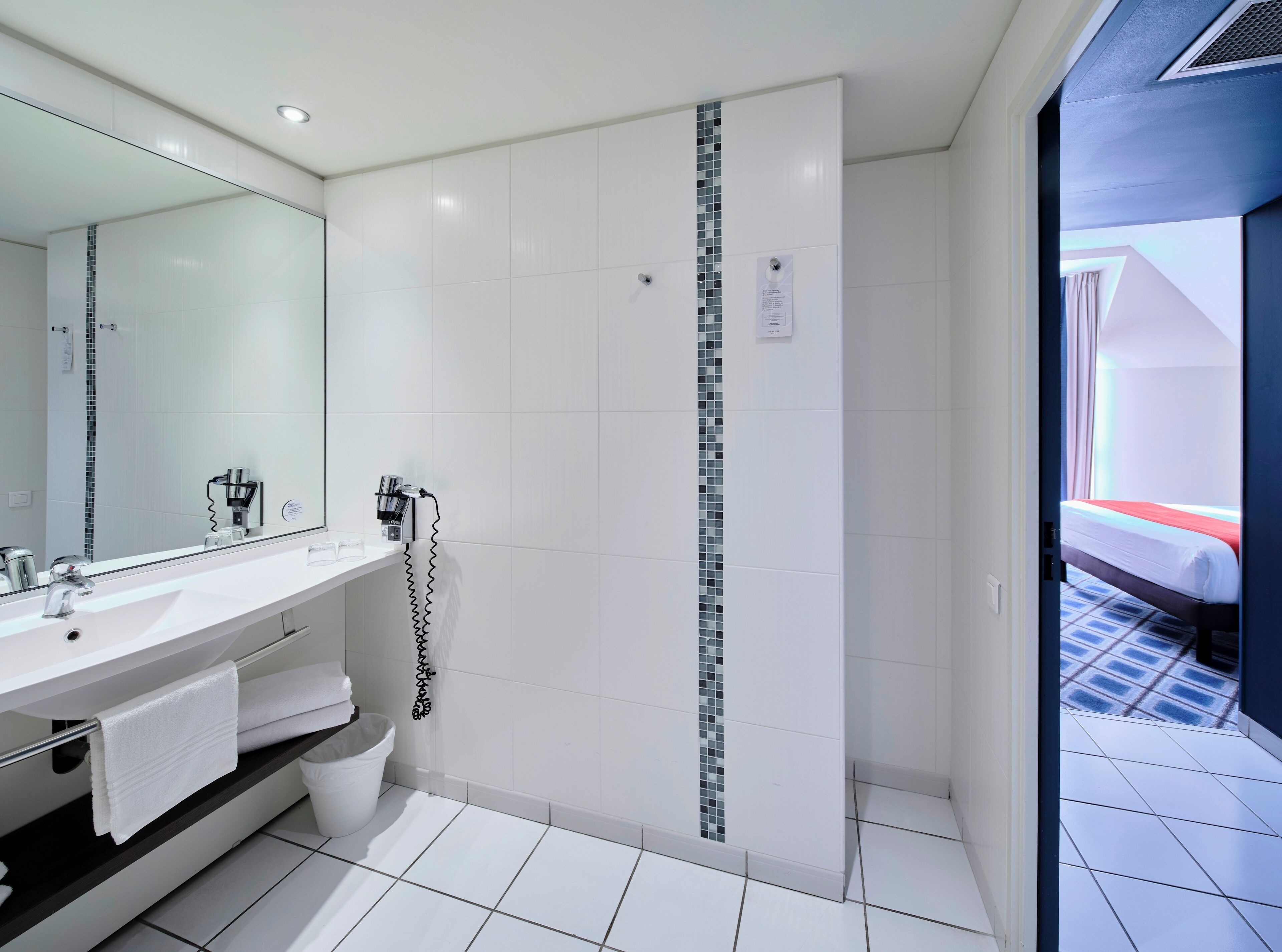 Een moderne badkamer met een grote spiegel, witte tegels, een douche en een bed in de aangrenzende kamer.