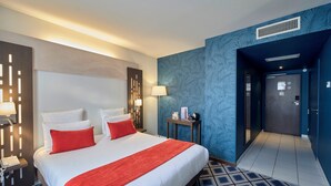 Habitación de hotel con una cama grande, dos almohadas rojas, una colcha blanca, un plaid rojo, una mesita de noche con lámpara, una pequeña mesa con lámpara y una pared con estampado azul.