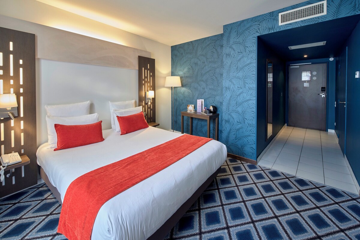 Une chambre d’hôtel avec un grand lit, deux oreillers rouges, un couvre-lit blanc, un plaid rouge, une table de chevet avec une lampe, une petite table avec une lampe et un mur à motifs bleus.