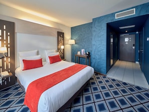 A hotel room with a large bed, two red pillows, a white bedspread, a red throw blanket, a nightstand with a lamp, a small table with a lamp, and a blue patterned wall.