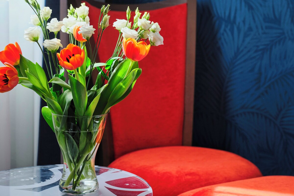 Une pièce avec un vase en verre rempli de fleurs posé sur une table, une chaise rouge et un mur bleu à motifs.