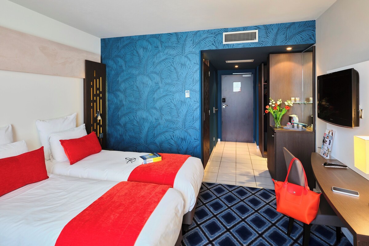 Une chambre d’hôtel avec deux lits, un bureau avec une chaise, une télévision et une salle de bain visible à travers une porte ouverte.
