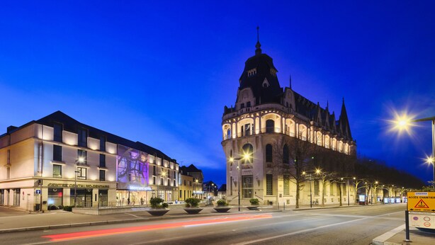 A historic building with a clock tower, adjacent to modern buildings with illuminated signage. - Mercure Chartres Centre Cathédrale (Chartres)