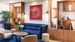 A modern lounge area with a blue sofa, round coffee table, and two armchairs. There is a potted plant, a framed artwork, and wooden shelves with decorative items. - Mercure Chartres Centre Cathédrale (Chartres)