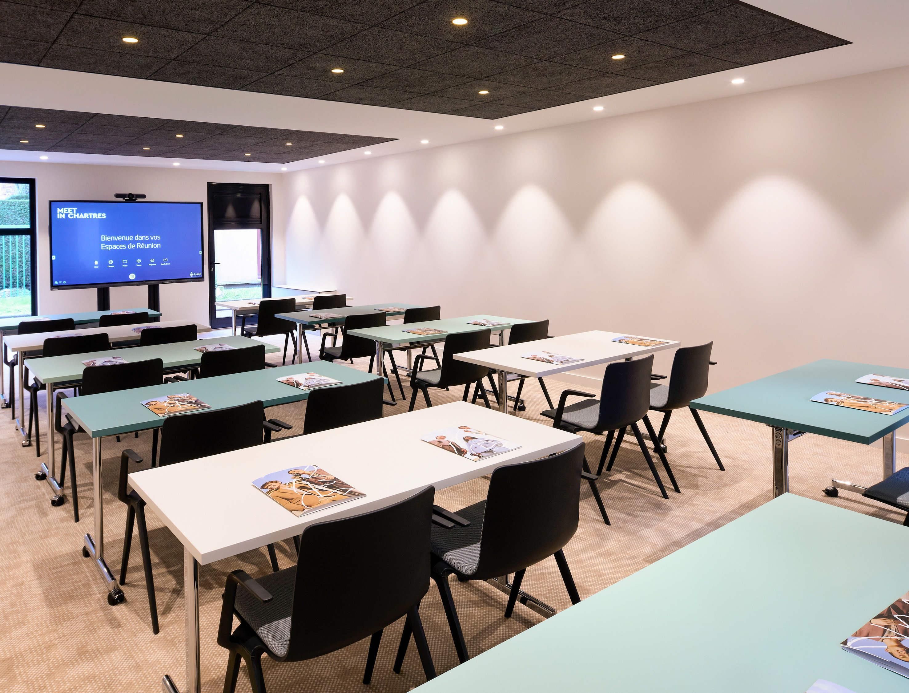 Une salle de conférence équipée de tables et de chaises, d’un écran de projection et d’une grande fenêtre.