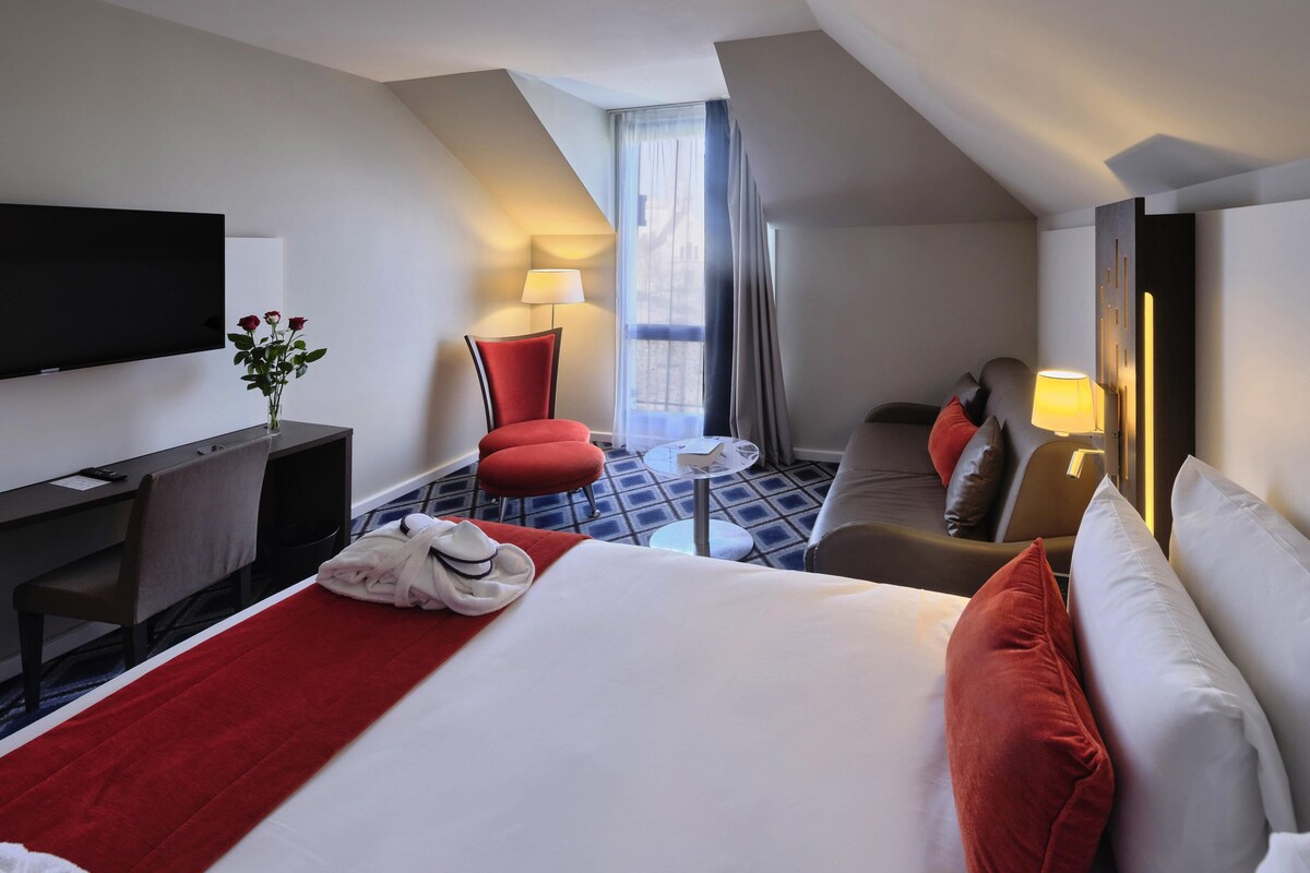 A hotel room with a bed, a desk, a TV, a chair, a sofa, and a window with curtains.