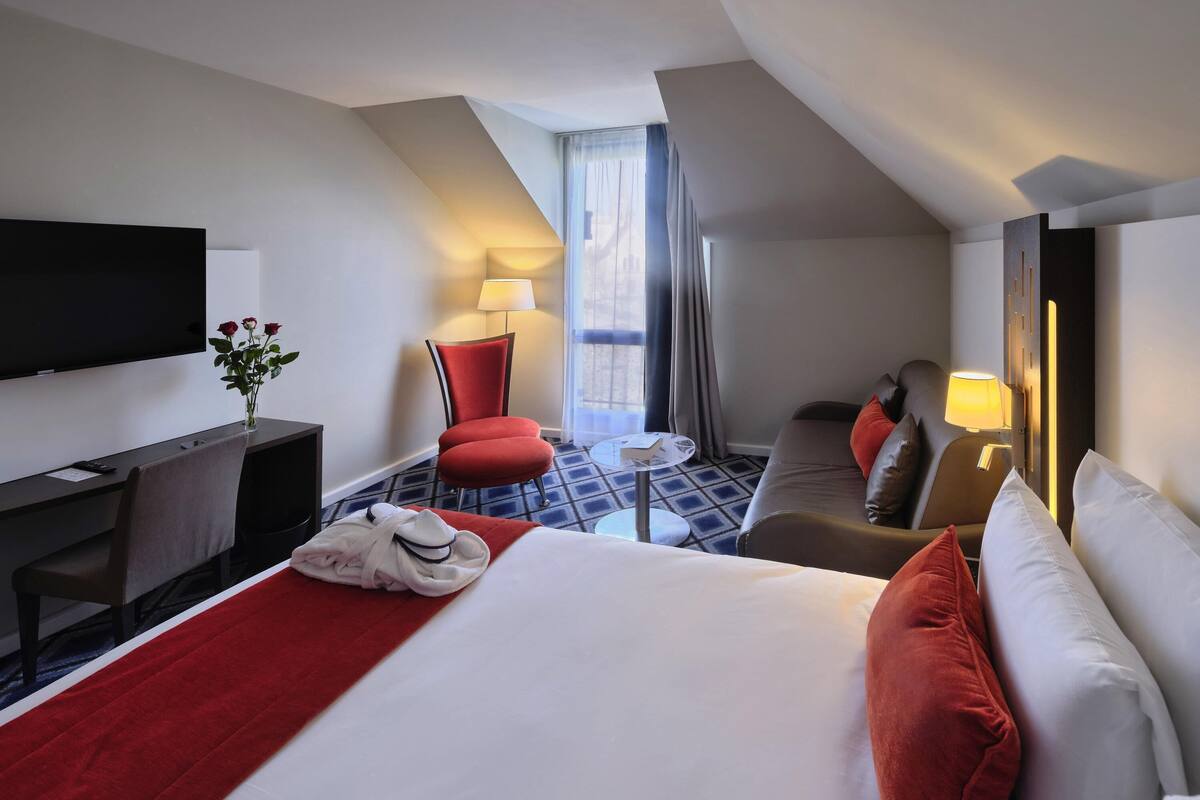 Ein Hotelzimmer mit einem Bett, einem Schreibtisch, einem Fernseher, einem Sessel, einer Couch und einem Fenster mit Vorhängen.