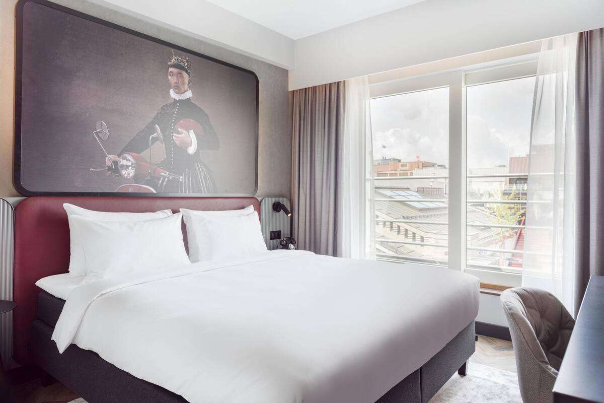 A modern hotel room with a large bed, a desk, and a large window offering a city view.