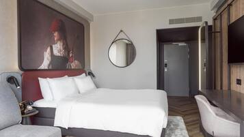 A modern hotel room with a large bed, a desk, a chair, a round mirror, and a painting on the wall.