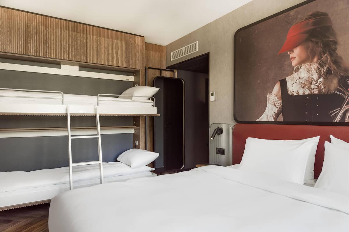 A hotel room with bunk beds, a large bed, a painting on the wall, and a radiator.