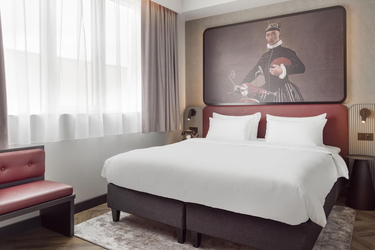 A modern hotel room with a large bed, a red upholstered bench, a large framed artwork on the wall, and a window with sheer curtains.