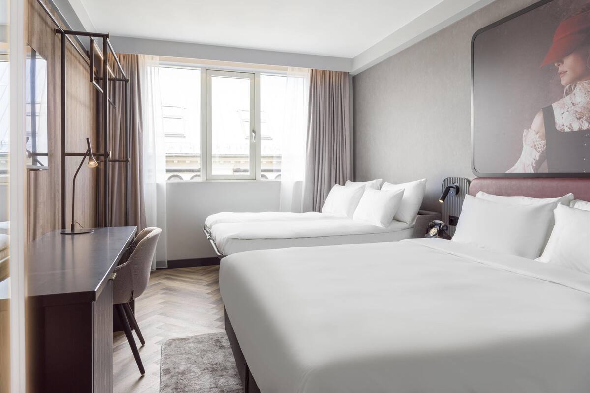 A hotel room with two beds, a desk, a chair, a lamp, and a large window with curtains.