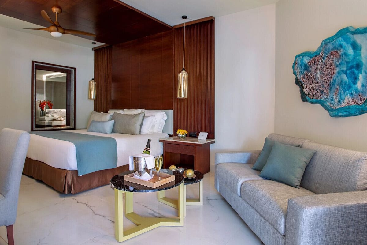 A hotel room with a bed, a sofa, a small table with a bottle and glasses, a mirror, and a decorative wall piece.