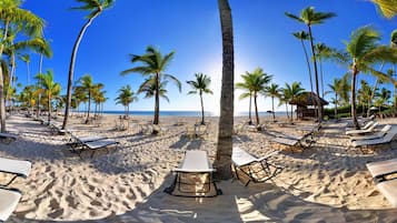 A beach with rows of sun loungers and palm trees, a thatched-roof structure, and a clear view of the ocean.