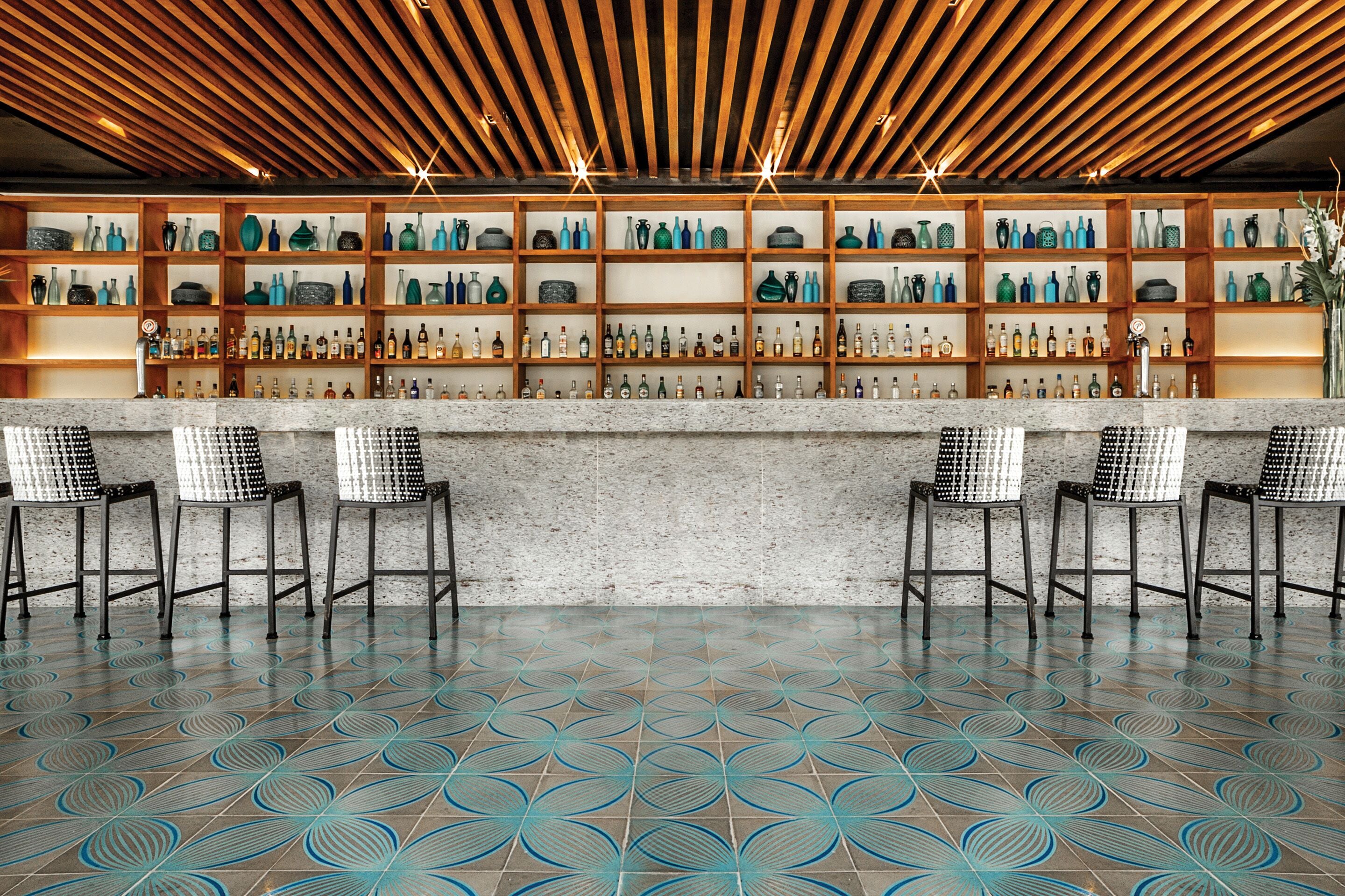 Zona de bar con piso estampado, repisas de madera con botellas y sillas blancas y negras.